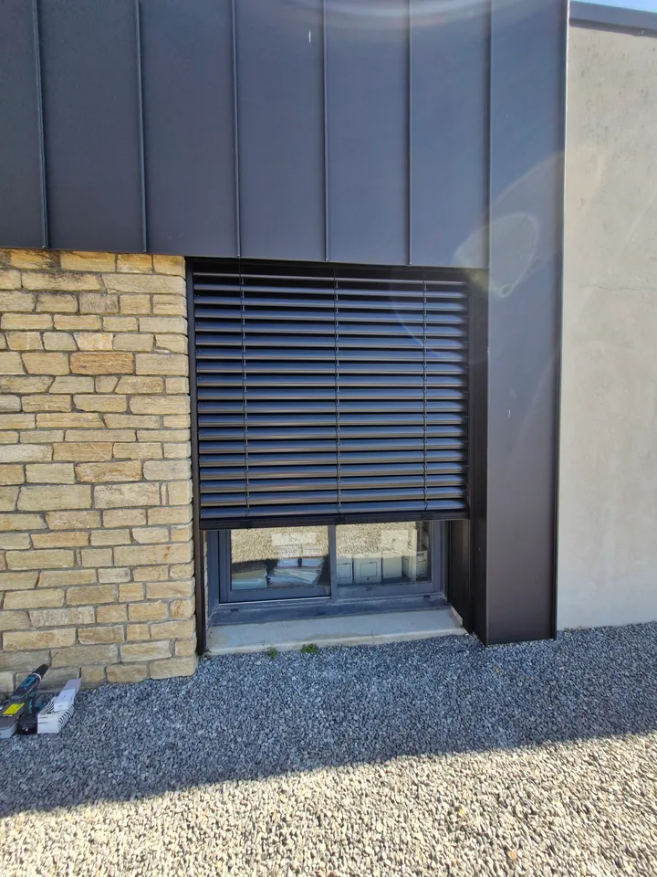 Brise-soleil orientable gris devant une ouverture en façade pierre et métal