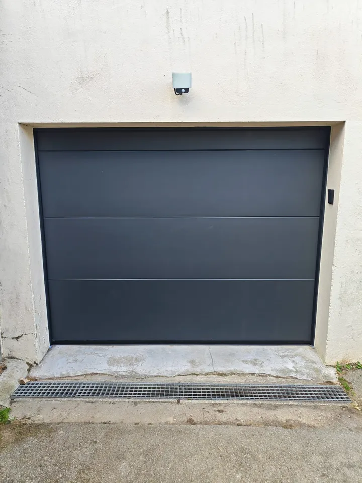 Porte de garage grise à larges panneaux dans un encadrement enduit